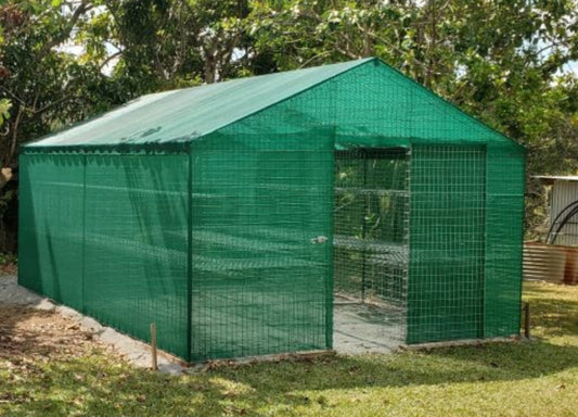 A green garden shadehouse made of  galvanised metal covered with shadecloth, situated outdoors among plants and trees.